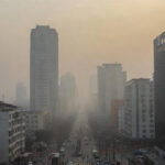 प्रदूषण से ढका एक बड़ा शहर, धुंध और स्मॉग में घिरी इमारतें और मास्क पहने चलते लोग।